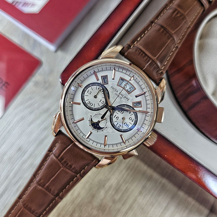Réplica AAA de reloj Patek Philippe en caja de oro rosa con esfera plateada texturizada, subdiales de cronógrafo, fecha y correa de cuero marrón.